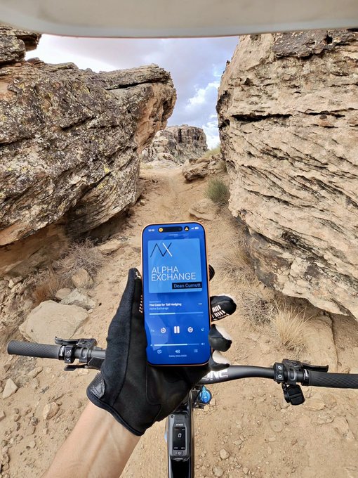 A first-person view of someone wearing black gloves riding a bicycle through a rocky canyon, with a smartphone mounted on the handlebars playing a song titled 'The Clock' from the 'Alpha Exchange' playlist.