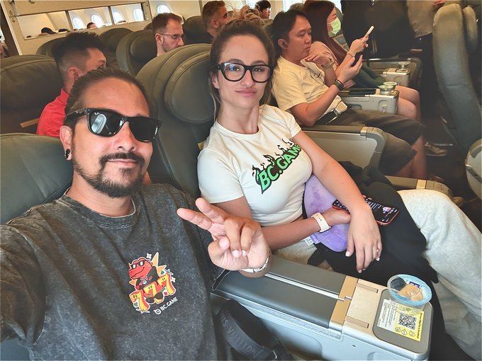 A man in sunglasses and a 'BC.GAME' t-shirt sits next to a woman wearing glasses and a 'BC.GAME' t-shirt on an airplane, taking a selfie.