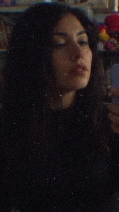 A dimly lit selfie of a young woman with long dark hair wearing a black top, taken in what appears to be a room with colorful items in the background.