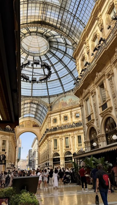 A grand shopping arcade featuring an ornate glass domed ceiling, with luxury storefronts including Louis Vuitton visible along the right side as shoppers walk through the polished marble floors.