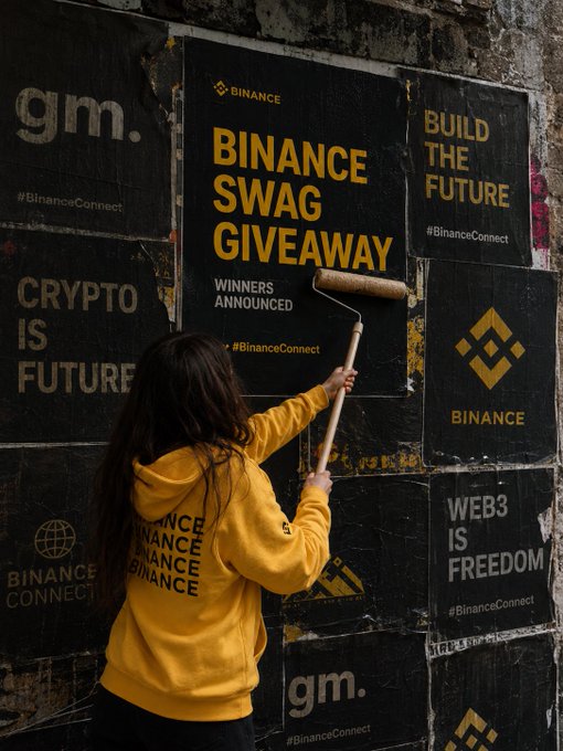 A woman in a yellow hoodie paints over Binance posters on a wall, with visible text including "BINANCE SWAG GIVEAWAY", "BUILD THE FUTURE", "WEB3 IS FREEDOM", and "gm".