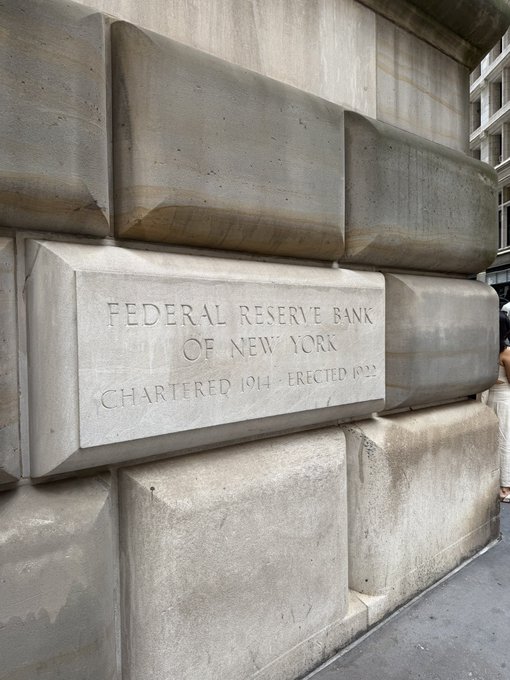 A close-up of a stone plaque engraved with 'FEDERAL RESERVE BANK OF NEW YORK, CHARTERED 1914, ERECTED 1922' set into the large block-like stone facade of the building.