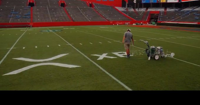 A worker uses a paint machine to mark "XRP" on a football field, referencing the recent X (formerly Twitter) rebrand and the accompanying social media post celebrating the ticker.