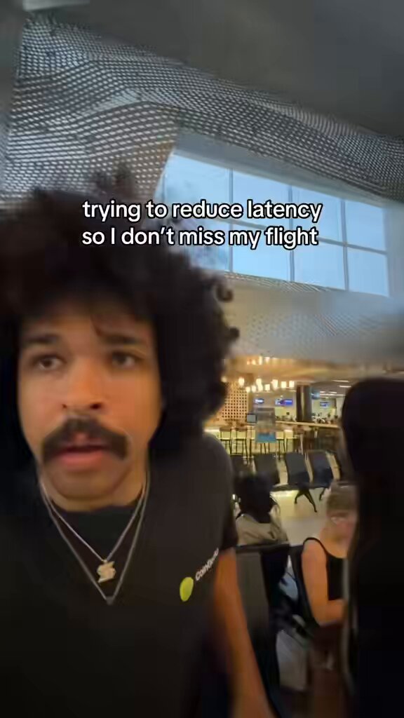 The image features a man with curly hair wearing a black shirt, who appears to be making a face or showing frustration while looking at his phone screen. The caption of the image reads "Trying to reduce lateness so I don't miss my flight". This suggests that the man is experiencing some sort of delay or inconvenience and is trying to communicate this issue through text on his phone.