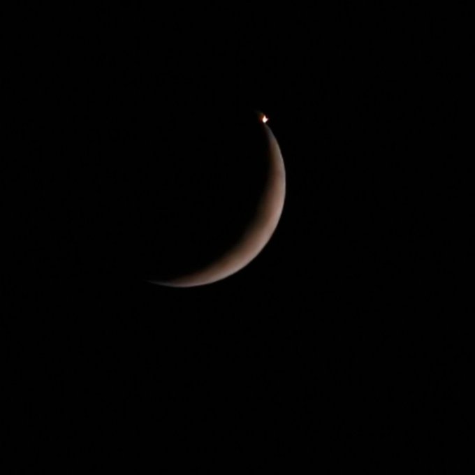 A crescent moon visible against a dark night sky, with a bright point of light (likely Venus) glowing at its upper tip.