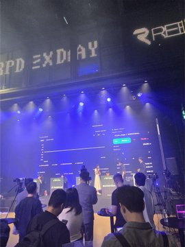 An audience watching a stage presentation labeled 'RED EXDAY' at what appears to be a tech or gaming conference, with presenters standing before large display screens showing data.