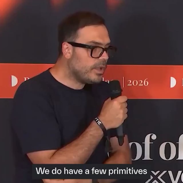 A man with glasses and a dark t-shirt speaks into a microphone on a stage with a red banner displaying '2026'; a subtitle reads 'We do have a few primitives'.
