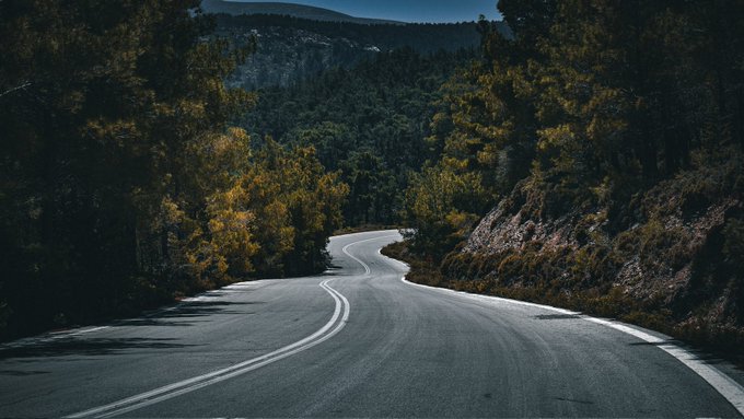 A scenic photograph of a winding asphalt road cutting through a dense forest with dark green trees and mountains in the background.