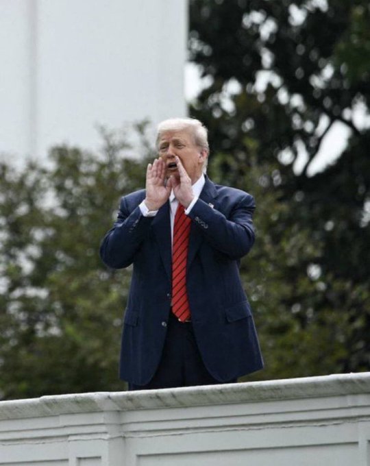 A man with blonde hair in a navy blue suit and red tie stands behind a white railing, calling out with both hands cupped around his mouth, with trees visible in the background.