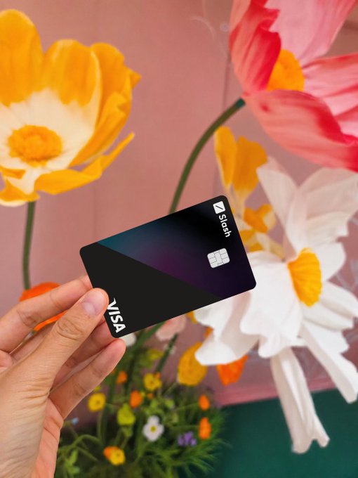 A hand holds a black Visa Slash card in front of a colorful floral arrangement and a painted wall.