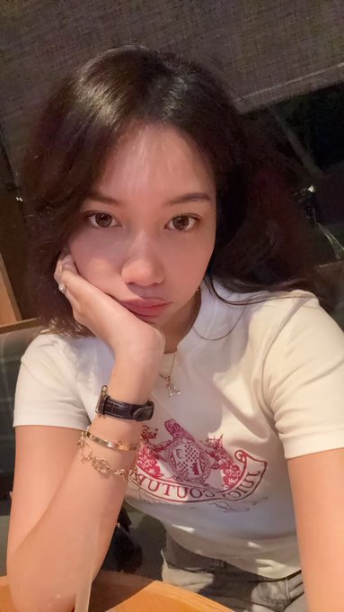 A young woman with long dark hair poses for a selfie with her hand against her cheek, wearing a white t-shirt featuring a red crest design with the text 'GUTUO 1878' visible.