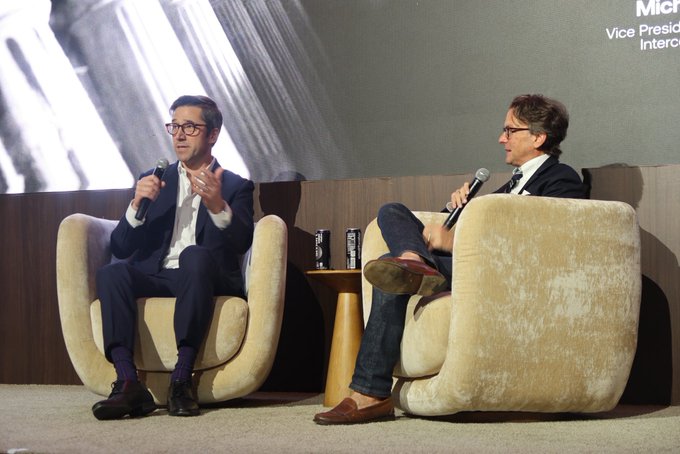 Two men are seated in plush, cream-colored armchairs on a stage during a panel discussion, each holding a microphone and speaking, with a large screen behind them partially displaying the title 'Vice Presi' and 'Interc'.