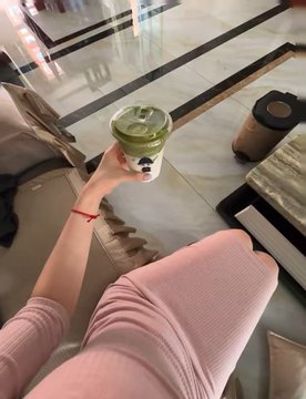 An overhead selfie of a person holding a green matcha latte in a white cup while sitting down wearing pink pants.
