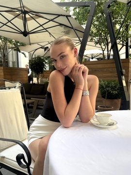 A young woman in a black top and white skirt sits at an outdoor cafe table under an umbrella, resting her chin on her hands next to a teacup.