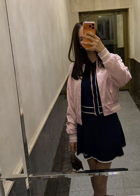 A woman in a pink bomber jacket and navy dress takes a mirror selfie in a hallway with her orange phone, captioning her commute to work where she's planning to browse for new purchases, explore NFT suggestions like Robin Hood NFTs, and find streams to connect with new people.