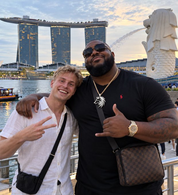 Two men posing together at Marina Bay Sands in Singapore with the iconic three-tower hotel visible in the background; one wears a white shirt making a peace sign, the other a large man in a black shirt with a lion pendant and designer bag.