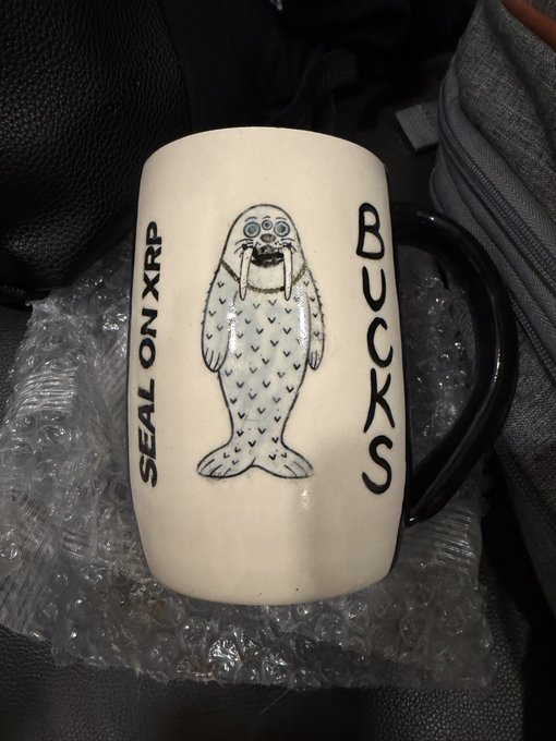 A white ceramic mug with black handle features a hand-drawn seal illustration and the text 'SEAL ON XRP' and 'BUCKS', sitting on bubble wrap.