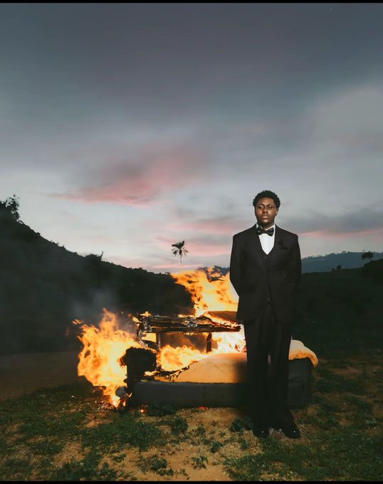 A cinematic photograph of a young man in a black tuxedo standing calmly beside a burning couch outdoors at dusk, with dramatic smoke and a pinkish sunset sky.