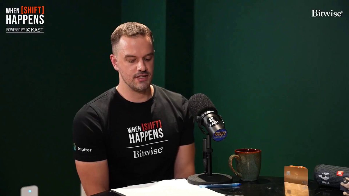 The image features a man sitting at a desk with a microphone, wearing a black t-shirt with white text. The background of the image shows a green wall, and there are two cups placed nearby - one near the top right corner and another towards the bottom left corner. The caption "When it happens" is written on the man's shirt, which suggests that he might be discussing the current state or trends in cryptocurrency markets.
