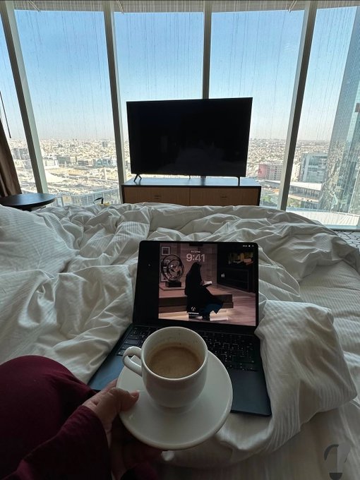 A first-person view from a hotel bed showing a hand holding a cappuccino, with a MacBook displaying a 9:41 lock screen in the foreground and a cityscape visible through large windows.