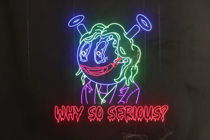 A neon sign depicting a stylized cartoon face with haloed headphones above and the glowing red text 'WHY SO SERIOUS?'