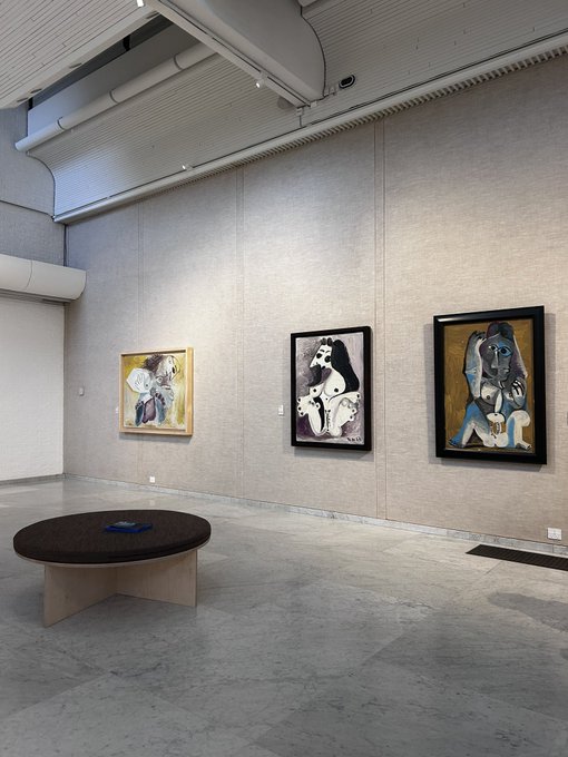 An art gallery interior showcases three framed Picasso paintings on a beige wall, with a round wooden table in the foreground on a polished concrete floor.