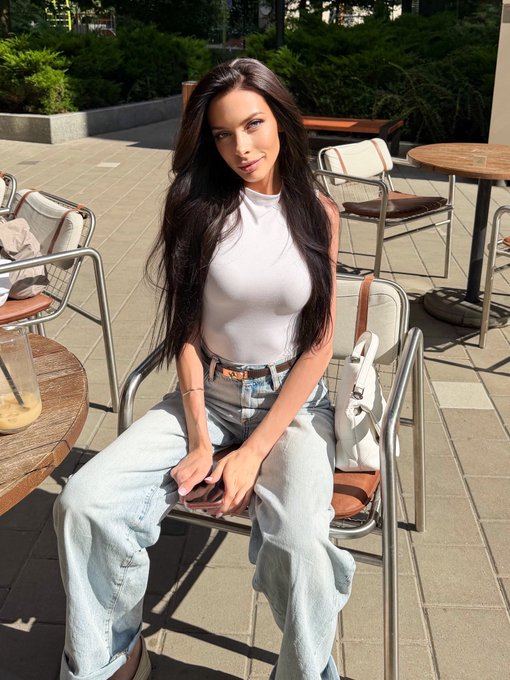 A woman with long dark hair sits at an outdoor café patio, wearing a white sleeveless top and light blue jeans, holding a phone with a white bag resting on the chair beside her.