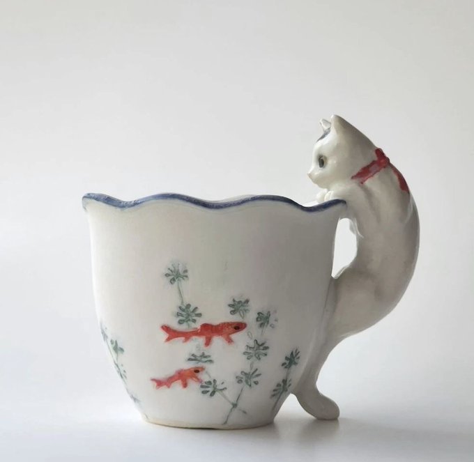 A white porcelain teacup with a scalloped blue rim and handle shaped like a cat's body and head, decorated with painted red fish and green floral motifs.