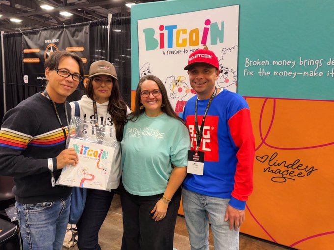 Four people pose in front of a 'BITCOIN: A Treasure to Hoard' banner with a tagline about fixing broken money, at what appears to be a convention booth.