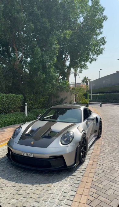 A silver and black Porsche sports car with a Dubai license plate reading '16197' is parked on a paved area beside a hedge and trees.
