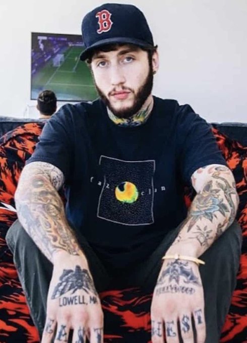 A heavily tattooed young man wearing a dark cap with the Boston Red Sox "B" logo and a black t-shirt sits on a red and black patterned couch, with "LOWELL, MA" and "BEST" visible on his hand tattoos. The image is tied to a report about FaZe Banks and Trader Rowdy investing early in the FOMO crypto presale, which has since grown 20x to $600M in yearly revenue.
