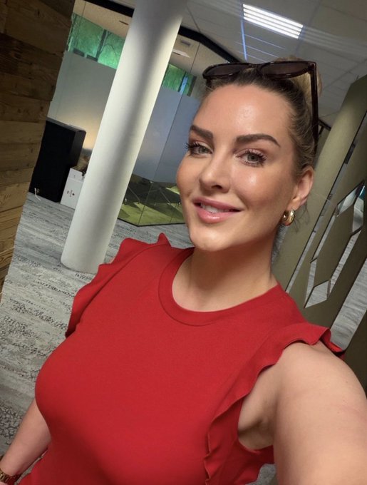 A woman in a red ruffled top smiles for a selfie in what appears to be a modern office space, with sunglasses perched on her head, perfectly capturing the cheerful "GM GM TGIF" (good morning, thank God it's Friday) vibe to start the day.