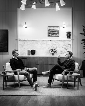 Two men sit in modern armchairs across from each other in a sleek, mid-century styled room, engaged in what appears to be an in-depth podcast or interview conversation. The image visually represents the long-awaited discussion between Micky Malka and David Senra, with the "$TIBBIR" ticker hinting at Ribbit Capital's involvement in the conversation topic.