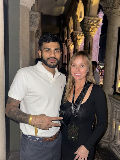 A photo of a man and a woman posing together in an ornate venue with carved stone columns; the man wears a white polo and has a tattooed arm, while the woman wears a black top and a lanyard.