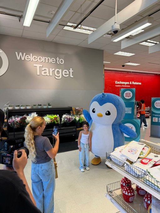 A young girl poses with a blue penguin mascot at a Target store entrance, where a woman with blonde hair takes a photo with her phone.