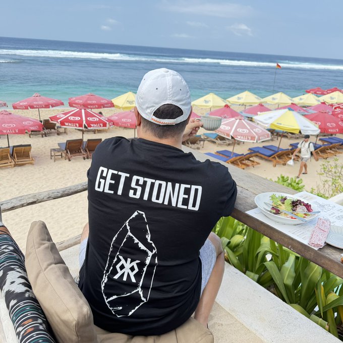 A man seen from behind wearing a white cap and a black t-shirt with the text 'GET STONED' and a Bitcoin logo, sitting at a beachside restaurant overlooking the ocean and colorful umbrellas.