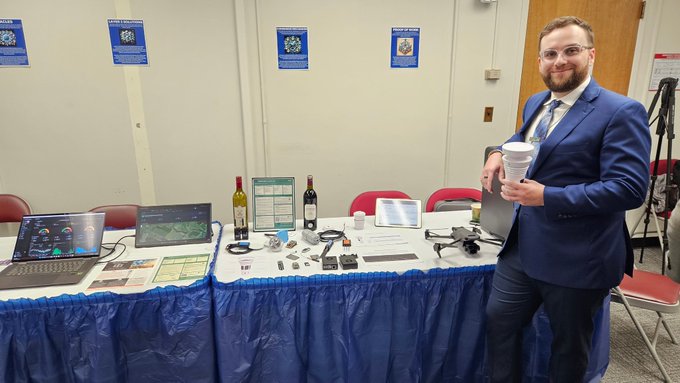 The image features a man standing next to a table displaying various items, including wine bottles and other objects such as a laptop and a cup of coffee. The man appears to be at a conference or event where he is showcasing innovative applications related to agriculture and insurance. The setting suggests that this gathering could have been organized by the New York State Bar Association (NYSBA) for Blockchain Demo Day, which was held in Albany on September 14th.