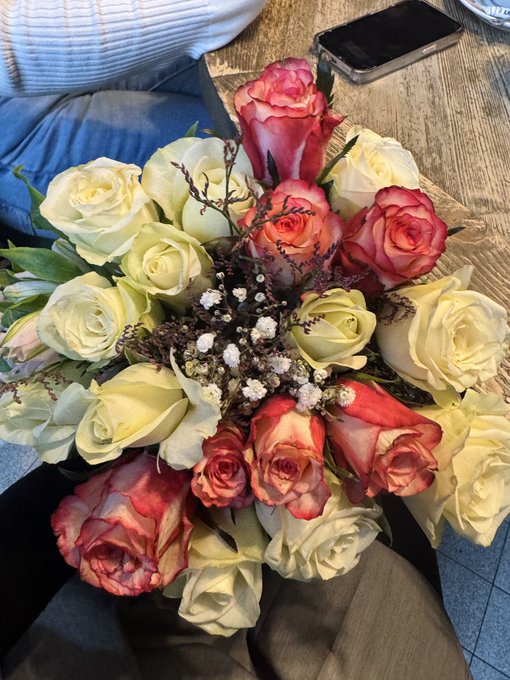 A bouquet of cream, pink, and red roses with lavender accents resting on a person's lap next to a wooden table with a smartphone.