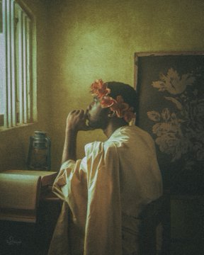 The image features a person wearing a white robe, with red flowers adorning their head. They are seated at a desk or table, which has an open book placed next to them. The background of the image includes a window that allows natural light into the room and a painting hanging on the wall behind the person.