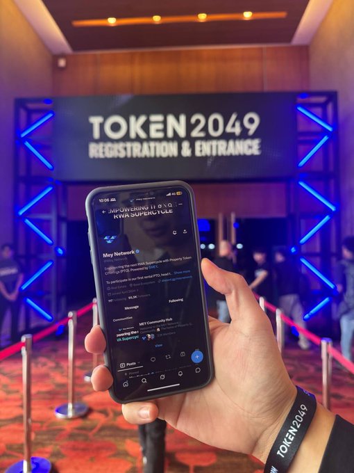 A hand holds a smartphone displaying a Twitter/X feed at the TOKEN 2049 conference registration and entrance, with the attendee wearing a TOKEN2049 wristband.
