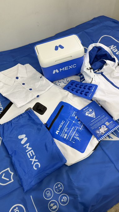 An overhead shot of various MEXC cryptocurrency exchange branded merchandise laid out on a blue surface, including a small cooler, white polo shirts, a drawstring bag, stickers, and informational flyers.