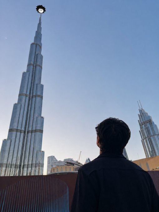 A person viewed from behind looks up at the Burj Khalifa skyscraper, which has a cute cartoon face sticker placed on its spire.