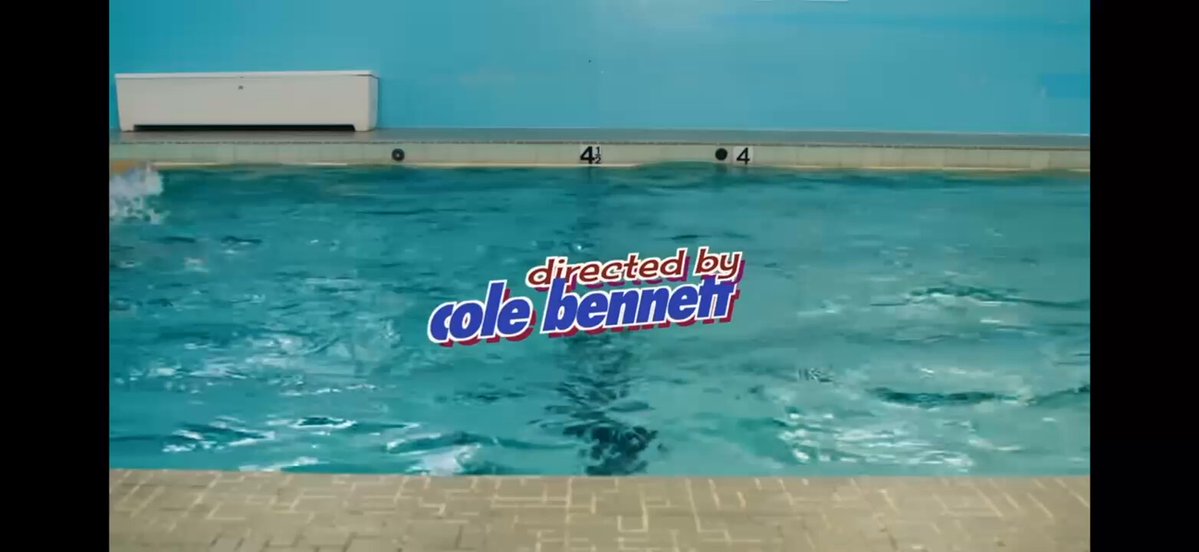 The image features a swimming pool filled with water, surrounded by a brick wall. The pool has blue tiles surrounding its edge, creating an inviting atmosphere for swimmers. In the background of the image, there are two people standing near the pool area, possibly enjoying their time in the water or taking a break from swimming.