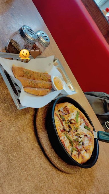 The image features a delicious pizza sitting on top of a black pan, accompanied by some breadsticks. The pizza has various toppings such as pepperoni, mushrooms, and onions. A small container filled with sauce can be seen next to the pizza. In addition to these items, there are two bottles placed nearby, one near the pizza and another further away from it. The background of the image shows a dining table surrounded by chairs, suggesting that this meal is being enjoyed in a restaurant or at home.