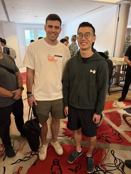 The image shows two smiling men posing together at what appears to be a casual networking event, with one wearing a white t-shirt and the other in a dark hoodie and glasses, standing on a red carpet with event attendees and decor visible in the background. This relates to the post about meeting people in person at the @hypurr_co event in Singapore, capturing a moment of the in-person connections the author enjoyed during the gathering.