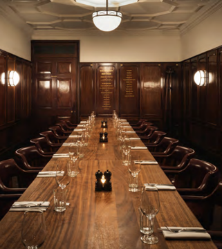 A long wooden dining table set with water glasses, candles, and folded napkins stretches down an elegant wood-paneled room with leather chairs and a pendant light overhead.