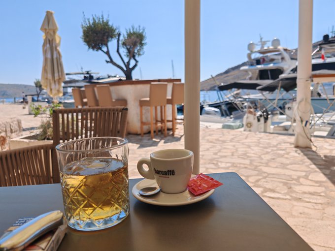 A glass of whiskey and an espresso cup branded 'Barcaffè' sit on a table at a waterfront café with yachts and a tree visible in the sunny background.