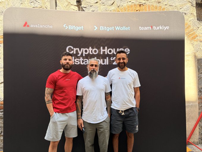 The image features three men standing next to each other, posing for a photo against a backdrop of a black banner with white text that says "Crypto House Istanbul". The men are wearing casual attire, including t-shirts and shorts. One man is holding a cell phone in his hand.