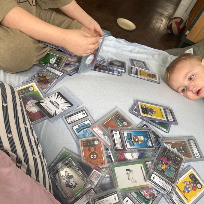 A young child lying next to a large collection of graded Pokémon trading cards spread out on a bed, with an adult's hand visible organizing the cards.