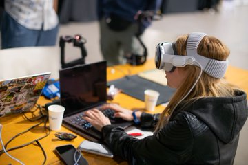 A young woman wearing a white Apple Vision Pro headset is focused on her laptop while working at a table scattered with cups, cables, and tech gear, capturing the late-night hacking energy of ETHGlobal Lisbon. The scene perfectly embodies the "the party is just getting started" spirit of Day 1 builders continuing their work deep into the evening.